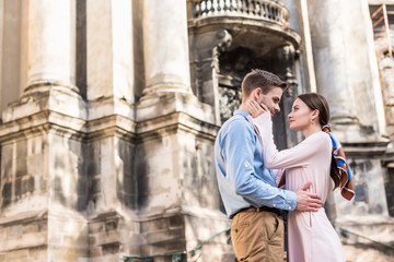beautiful young woman touching face of happy boyfriend while standing near ancient building in sunlight