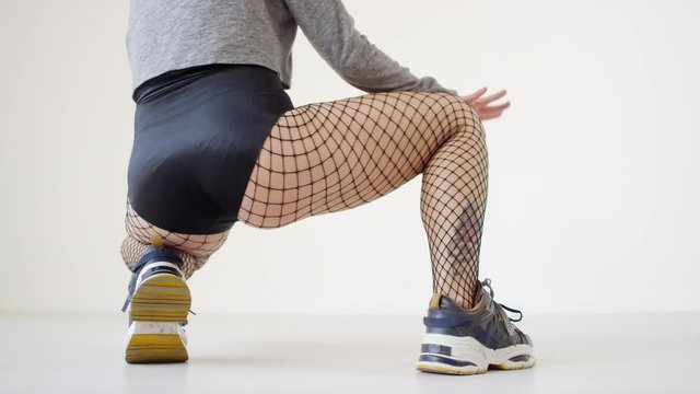 Low section rear view shot of sexy female dancer in fishnet tights and shorts getting into squat, shaking her buttocks and thrusting hips while twerking in studio