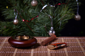 An ashtray with a cigar and a lighter on the table surface against the background of the Christmas tree.