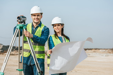 Smiling surveyors with digital level and blueprint looking at camera
