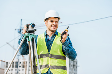 Smiling surveyor with digital level talking on radio set with construction site on background