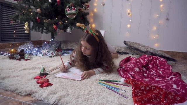 little girl with a rim of a deer colorizes a picture of Santa Claus. Changes the color of the pencils. The child lies under the tree.