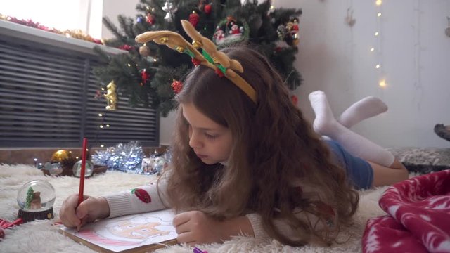 Curly girl with deer horns draws a picture of Santa Claus. The girl lies on the back of the Christmas tree and draws in red pencil. Approaches the camera to the girl. New Year's mood.