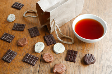 Handmade dark, white and milk chocolate, a cup of tea and paper packaging bag on a wooden table background. Top view