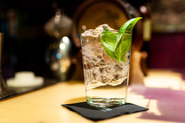 close up of gin tonic glass inside bar on top of bar counter with ice cubes and basil leaves at night