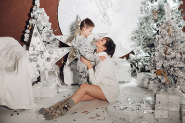 Fototapeta na wymiar Positive little cute boy with beautiful mom embracing in stylish decorated studio for Christmas holiday. Lovely mother holding her baby and smiling. Concept of New Year.