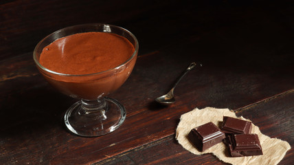 A bowl with chocolate mousse, pieces of dark chocolate and a teaspoon on the old dark brown table background