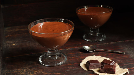 Chocolate mousse in transparent bowls, pieces of dark chocolate and a teaspoon on the old dark brown table background
