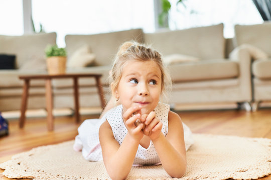 Charming cute little girl with chubby cheeks lying on carpet with sofa in background. Cute female child lying on floor in living room, holding hands under her chin, looking away with dreamy smile