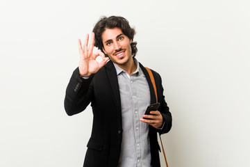 Young business man holding a phone cheerful and confident showing ok gesture.