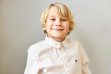 Adorable blonde European boy wearing stylish shirt looking at camera with joyful smile, being in good mood, expressing joy. Happy childhood and positive human facial expressions and emotions