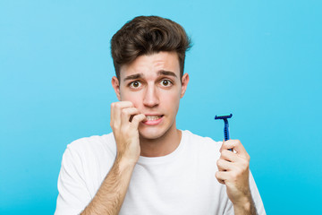 Young caucasian man holding a razor blade biting fingernails, nervous and very anxious.
