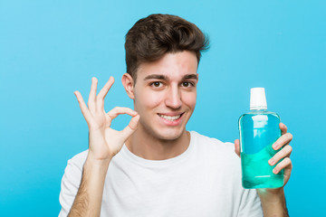 Young caucasian man holding a mouthwash cheerful and confident showing ok gesture.