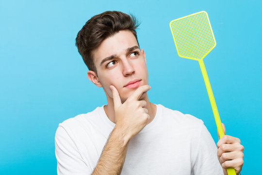Young caucasian man holding a fly swatter looking sideways with doubtful and skeptical expression.