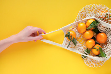 A human hand holds a string bag with tangerines on bold yellow color background.