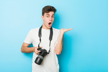 Young caucasian photographer man holding a camera impressed holding copy space on palm.