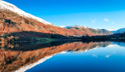 Fototapeta na wymiar Ullswater, Lake District with mountain reflections in the lake on a calm day with still waters. Snow capped mountains surrounding the second largest lake in the Cumbria, UK.