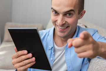 Attractive young man with e-book indicates happily at you, sitting on sofa at home, portrait, toned