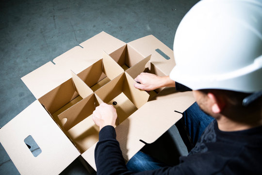 Man with a hard hat assembling a big, open, empty cardboard box with compartments on a stony floor in a warehouse.