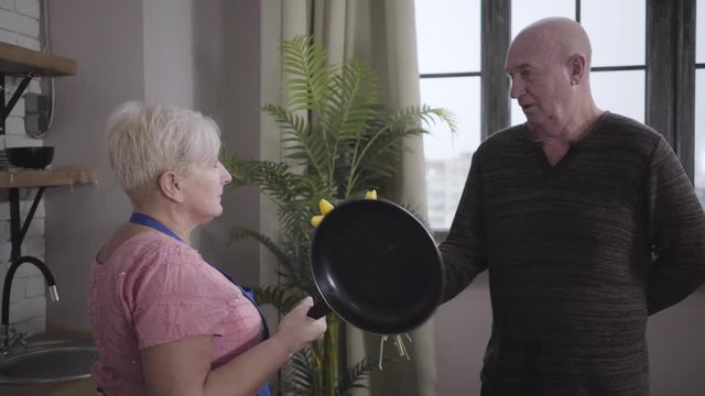 Portrait of mature Caucasian baldheaded man apologising with a bouquet of yellow flowers. Angry wife threatening him with frying pan. Wife educating her guilty spouse. Retirees having fun.