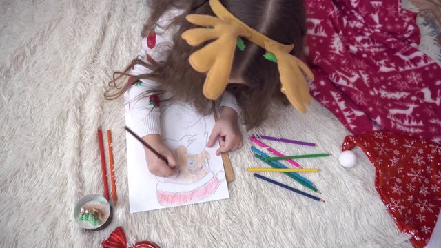 curly girl with deer horns paints a picture of Santa Claus. The girl lies on the back of the Christmas tree and draws with a brown pencil.shooting shit