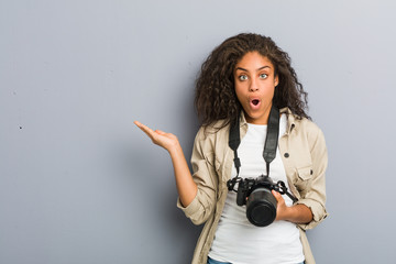 Young african american photographer woman holding a camera impressed holding copy space on palm.