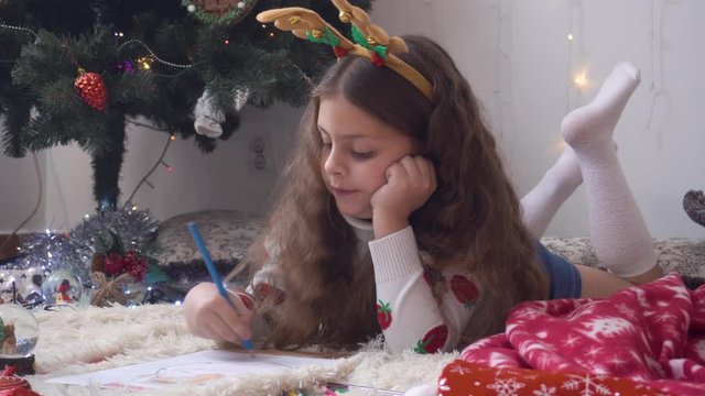 curly girl with deer horns paints a picture of Santa Claus. The girl lies on the back of the Christmas tree and draws with a blue pencil.