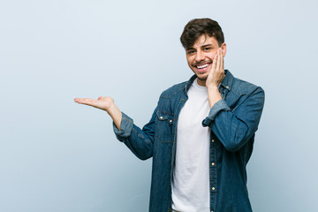 Young hispanic cool man holds copy space on a palm, keep hand over cheek. Amazed and delighted.