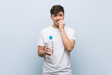 Young hispanic man holding a water bottle biting fingernails, nervous and very anxious.