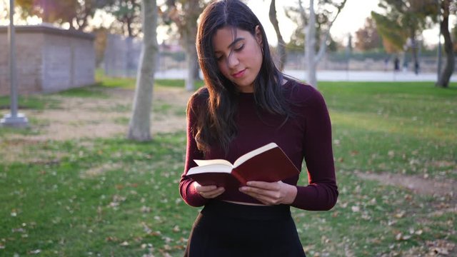 A young woman university student walking while reading a textbook outdoors in the college campus park at sunset SLOW MOTION.
