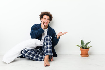 Mature man wearing pajama sitting on house floor holds copy space on a palm, keep hand over cheek. Amazed and delighted.