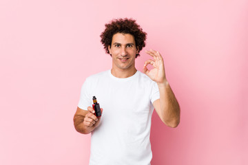Young caucasian curly man holding a vape cheerful and confident showing ok gesture.