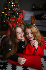 The concept of new year's house party. Large portraits of two redheads happy pretty girls girlfriends sisters in Christmas bright home sweaters laughing and celebrating Christmas.