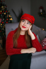 Beautiful bright attractive redhead girl plus size in a red sweater, beige leather skirt and red tights and a red cap rejoices and smiles on the background of the Christmas tree, gifts and bokeh.