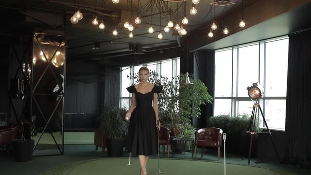 Full length stock video of stunning elegant lady in black evening dress walking towards the camera on artificial golf court with a golf stick and a ball. She is playing with a ball throwing it in the