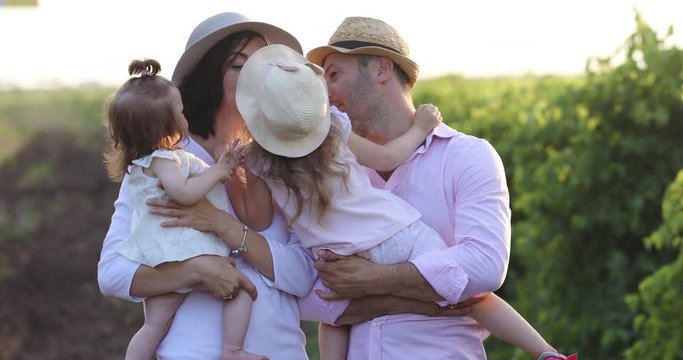 Young parents in hats kiss pretty children against against green vineyard at bright sunshine, summertime