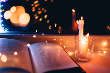 An open reading book lies on a window with glasses and light from a garland and candles....