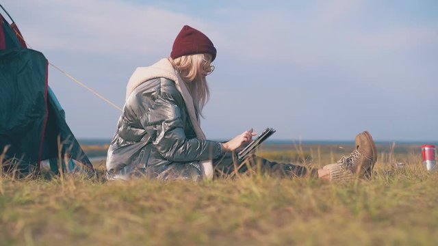 blonde haired lady in silver jacket with modern tablet sits by tent on green grass against clear sky at calm sunset