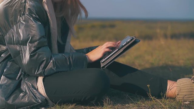 girl tourist in warm jacket works on modern tablet sitting on grass against clear sky at sunset close view