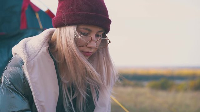 pretty girl tourist in stylish glasses and red knitted hat sits on river bank against clear sky at autumn sunset closeup