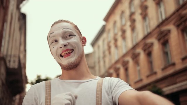 Smiling young mime is performing on street in white makeup. Performer in gloves follows somebody with eyes, waving, moving arms, winking, then angry with his hand. Facial expressions change quickly.