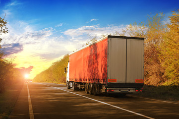 A truck and a trailer on the countryside road with an autumn forest against a sky with a sunset