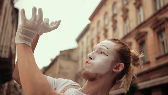 Handsome young mime is performing on street. Performer in white gloves pretends holding something invisible in hands, looking concentrated, surprised, then throws it over camera, satisfied, smiling.