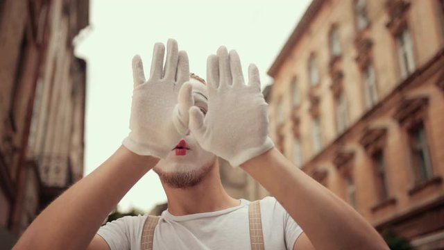 Handsome young mime is performing on street in special makeup. Performer in white gloves plays with camera, hiding behind hands, moving head to sides, down, looking satisfied, surprised, nodding.