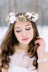 Naklejka premium Beautiful bride touching her curly hair, with floral head band wreath, made from flowers and cotton, standing outdoors, on the background of winter forest