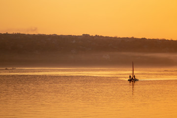 Fototapeta premium A sailboat sailing along the river in the light of evening sunset and a haze of fog creeping along the river.