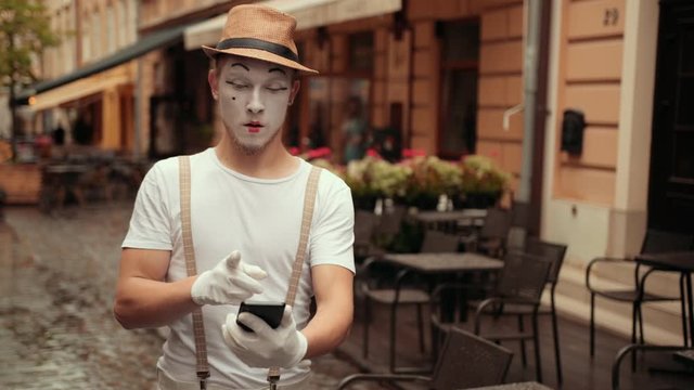 Handsome young mime with concentrated facial expression dials number on his phone. Performer taps on screen of cellphone, then puts near ear, calling.
