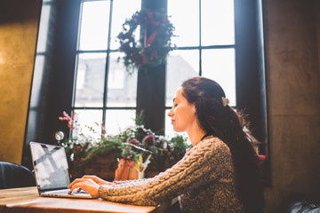 beautiful young brunette woman using a laptop at coffee shop at a wooden table near window typing...