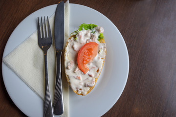Bread roll or bun with crab salad and tomato served with napkin and cutlery on a white plate on a brown table, copy space, high angle view from above