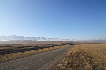 Asphalt road runs along the steppe to the snowy mountains.